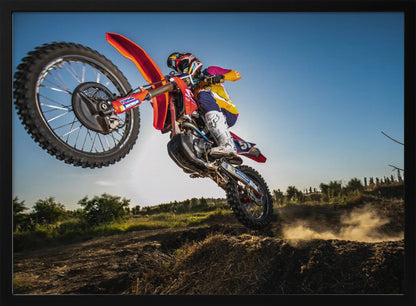 A dynamic, low-angle shot of a motocross rider in mid-air, jumping over a dirt mound. The rider is on a red, white, and blue dirt bike, wearing colorful racing gear and a helmet. The bike's front wheel is high in the air against a clear blue sky, while the back wheel is just clearing the jump, kicking up a cloud of dust. The background features a green, rural landscape. Wall Art
