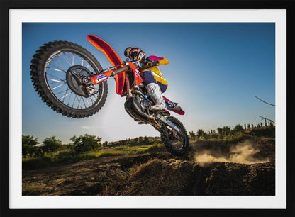 A dynamic, low-angle shot of a motocross rider in mid-air, jumping over a dirt mound. The rider is on a red, white, and blue dirt bike, wearing colorful racing gear and a helmet. The bike's front wheel is high in the air against a clear blue sky, while the back wheel is just clearing the jump, kicking up a cloud of dust. The background features a green, rural landscape. Wall Art