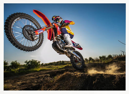 A dynamic, low-angle shot of a motocross rider in mid-air, jumping over a dirt mound. The rider is on a red, white, and blue dirt bike, wearing colorful racing gear and a helmet. The bike's front wheel is high in the air against a clear blue sky, while the back wheel is just clearing the jump, kicking up a cloud of dust. The background features a green, rural landscape. Wall Art
