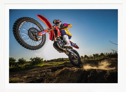 A dynamic, low-angle shot of a motocross rider in mid-air, jumping over a dirt mound. The rider is on a red, white, and blue dirt bike, wearing colorful racing gear and a helmet. The bike's front wheel is high in the air against a clear blue sky, while the back wheel is just clearing the jump, kicking up a cloud of dust. The background features a green, rural landscape. Wall Art