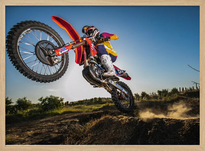 A dynamic, low-angle shot of a motocross rider in mid-air, jumping over a dirt mound. The rider is on a red, white, and blue dirt bike, wearing colorful racing gear and a helmet. The bike's front wheel is high in the air against a clear blue sky, while the back wheel is just clearing the jump, kicking up a cloud of dust. The background features a green, rural landscape. Wall Art