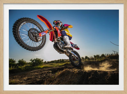 A dynamic, low-angle shot of a motocross rider in mid-air, jumping over a dirt mound. The rider is on a red, white, and blue dirt bike, wearing colorful racing gear and a helmet. The bike's front wheel is high in the air against a clear blue sky, while the back wheel is just clearing the jump, kicking up a cloud of dust. The background features a green, rural landscape. Wall Art