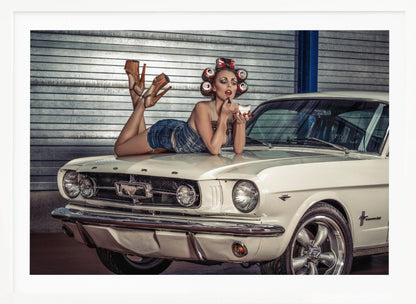 A woman in a retro pin-up style lies on the hood of a vintage white Ford Mustang in a garage, applying red lipstick with her hair set in soda can curlers. Poster