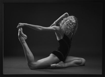 A black and white studio photograph of a flexible ballerina in a dark leotard and pointe shoes, arching her back to hold her extended leg over her head in an elegant pose against a dark background. Artwork