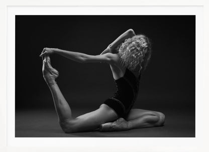 A black and white studio photograph of a flexible ballerina in a dark leotard and pointe shoes, arching her back to hold her extended leg over her head in an elegant pose against a dark background. Artwork