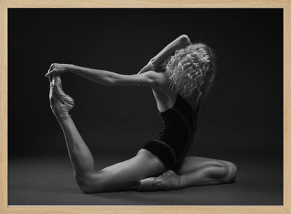A black and white studio photograph of a flexible ballerina in a dark leotard and pointe shoes, arching her back to hold her extended leg over her head in an elegant pose against a dark background. Artwork