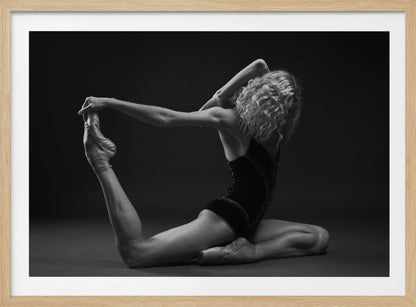 A black and white studio photograph of a flexible ballerina in a dark leotard and pointe shoes, arching her back to hold her extended leg over her head in an elegant pose against a dark background. Artwork