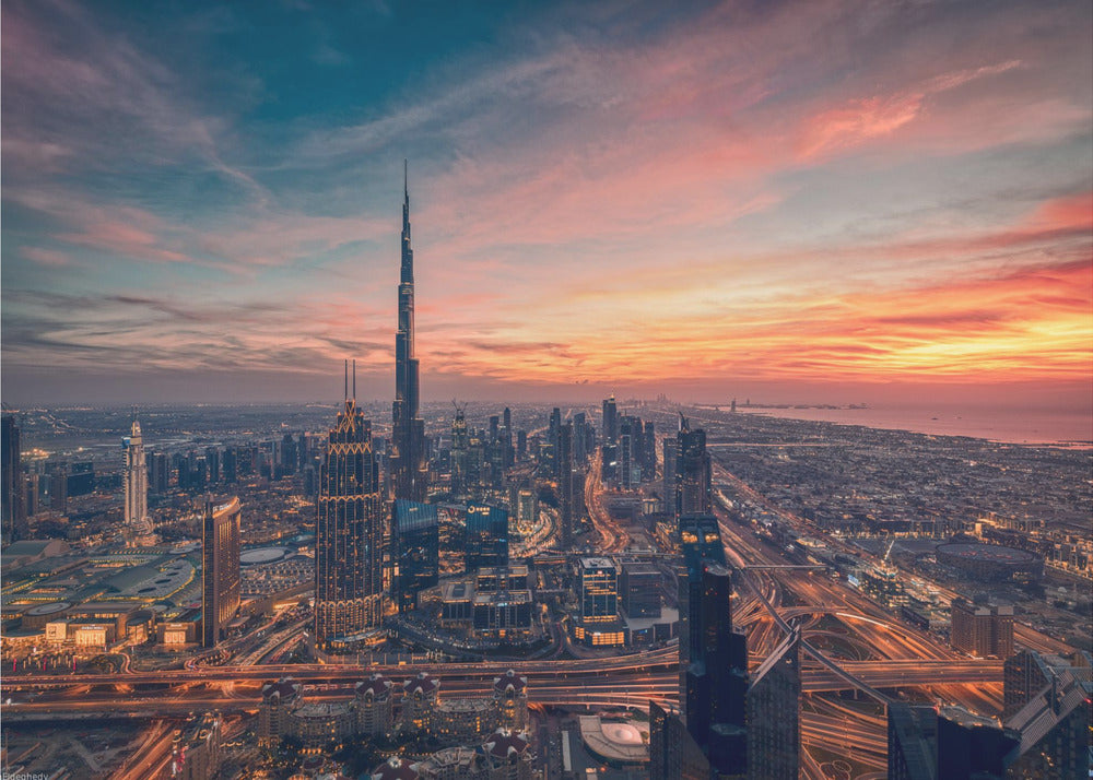 An aerial view of the Dubai skyline at sunset, with the Burj Khalifa prominently featured against a dramatic sky filled with orange, pink, and blue clouds. The sprawling city below is illuminated with golden lights from buildings and busy highways. Decor