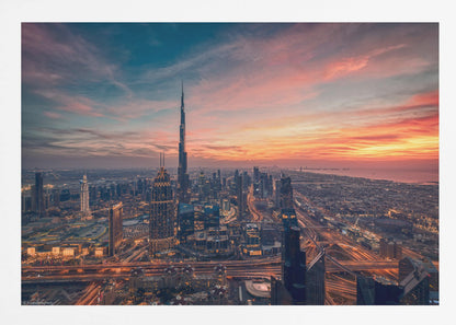 An aerial view of the Dubai skyline at sunset, with the Burj Khalifa prominently featured against a dramatic sky filled with orange, pink, and blue clouds. The sprawling city below is illuminated with golden lights from buildings and busy highways. Decor