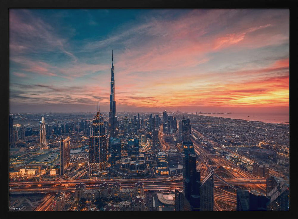 An aerial view of the Dubai skyline at sunset, with the Burj Khalifa prominently featured against a dramatic sky filled with orange, pink, and blue clouds. The sprawling city below is illuminated with golden lights from buildings and busy highways. Decor