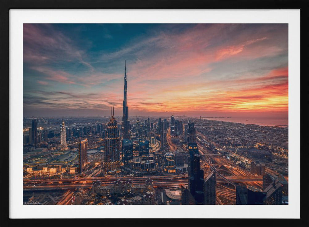 An aerial view of the Dubai skyline at sunset, with the Burj Khalifa prominently featured against a dramatic sky filled with orange, pink, and blue clouds. The sprawling city below is illuminated with golden lights from buildings and busy highways. Decor