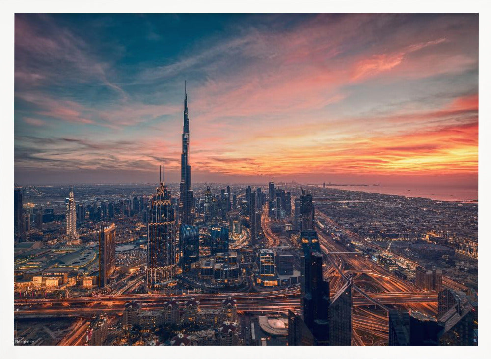 An aerial view of the Dubai skyline at sunset, with the Burj Khalifa prominently featured against a dramatic sky filled with orange, pink, and blue clouds. The sprawling city below is illuminated with golden lights from buildings and busy highways. Decor