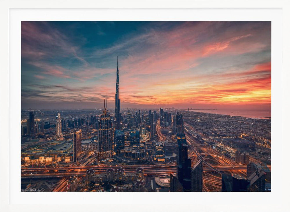 An aerial view of the Dubai skyline at sunset, with the Burj Khalifa prominently featured against a dramatic sky filled with orange, pink, and blue clouds. The sprawling city below is illuminated with golden lights from buildings and busy highways. Decor