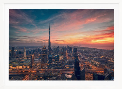 An aerial view of the Dubai skyline at sunset, with the Burj Khalifa prominently featured against a dramatic sky filled with orange, pink, and blue clouds. The sprawling city below is illuminated with golden lights from buildings and busy highways. Decor