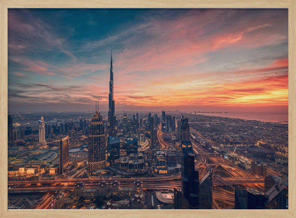 An aerial view of the Dubai skyline at sunset, with the Burj Khalifa prominently featured against a dramatic sky filled with orange, pink, and blue clouds. The sprawling city below is illuminated with golden lights from buildings and busy highways. Decor