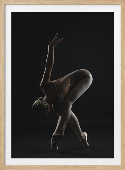 A dramatic studio photograph of a ballerina in a complex pose against a solid black background. Strong rim lighting highlights the graceful curves of her body as she bends forward with her legs crossed, one arm extended high and the other reaching towards her pointe shoes. Poster