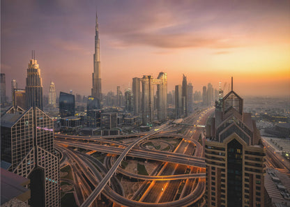 An elevated photograph of the Dubai skyline at sunset, featuring the towering Burj Khalifa against a hazy orange and purple sky. In the foreground, a complex network of illuminated highways and overpasses weaves between modern skyscrapers. Wall Art