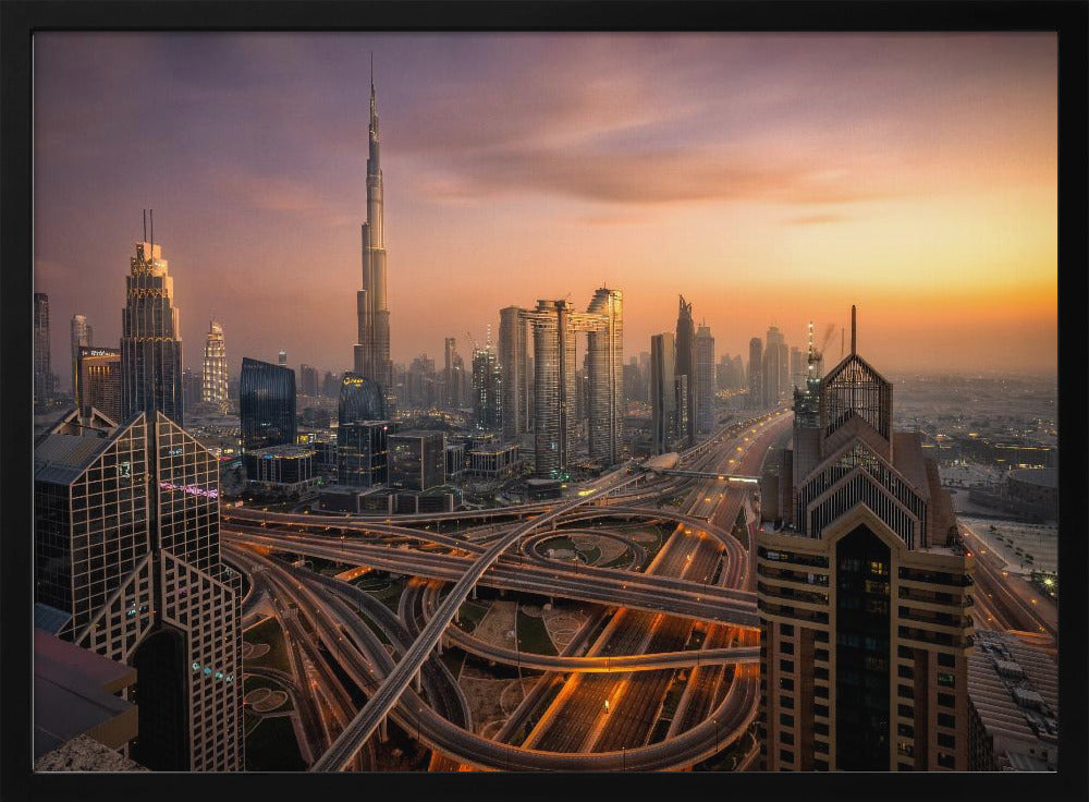 An elevated photograph of the Dubai skyline at sunset, featuring the towering Burj Khalifa against a hazy orange and purple sky. In the foreground, a complex network of illuminated highways and overpasses weaves between modern skyscrapers. Wall Art