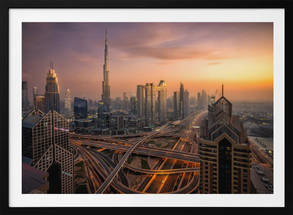 An elevated photograph of the Dubai skyline at sunset, featuring the towering Burj Khalifa against a hazy orange and purple sky. In the foreground, a complex network of illuminated highways and overpasses weaves between modern skyscrapers. Wall Art