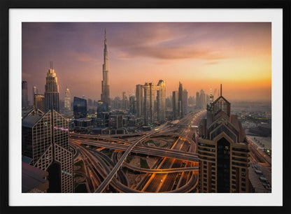 An elevated photograph of the Dubai skyline at sunset, featuring the towering Burj Khalifa against a hazy orange and purple sky. In the foreground, a complex network of illuminated highways and overpasses weaves between modern skyscrapers. Wall Art