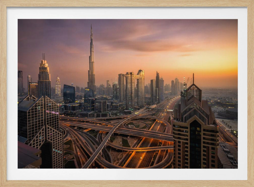 An elevated photograph of the Dubai skyline at sunset, featuring the towering Burj Khalifa against a hazy orange and purple sky. In the foreground, a complex network of illuminated highways and overpasses weaves between modern skyscrapers. Wall Art