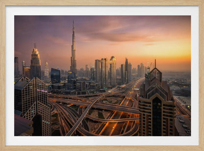 An elevated photograph of the Dubai skyline at sunset, featuring the towering Burj Khalifa against a hazy orange and purple sky. In the foreground, a complex network of illuminated highways and overpasses weaves between modern skyscrapers. Wall Art