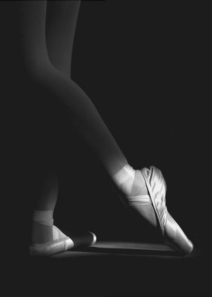 A dramatic black and white photograph showing the lower legs and feet of a ballet dancer, with one foot gracefully en pointe, highlighted by strong side lighting against a deep black background. Decor