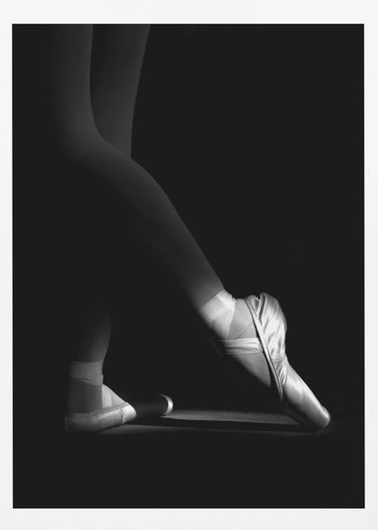 A dramatic black and white photograph showing the lower legs and feet of a ballet dancer, with one foot gracefully en pointe, highlighted by strong side lighting against a deep black background. Decor