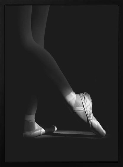 A dramatic black and white photograph showing the lower legs and feet of a ballet dancer, with one foot gracefully en pointe, highlighted by strong side lighting against a deep black background. Decor