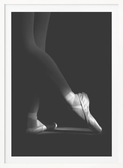 A dramatic black and white photograph showing the lower legs and feet of a ballet dancer, with one foot gracefully en pointe, highlighted by strong side lighting against a deep black background. Decor