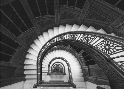 A mesmerizing black and white photograph shot from a high angle looking down into the center of a spiral staircase. The bright white marble steps contrast with the dark, ornate metal railing, creating a repeating arched pattern that draws the eye downward into a seemingly endless tunnel. The image is presented within a silver-colored frame. Artwork