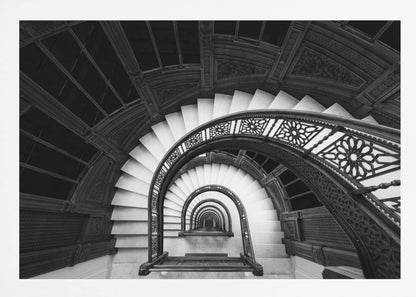 A mesmerizing black and white photograph shot from a high angle looking down into the center of a spiral staircase. The bright white marble steps contrast with the dark, ornate metal railing, creating a repeating arched pattern that draws the eye downward into a seemingly endless tunnel. The image is presented within a silver-colored frame. Artwork