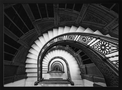 A mesmerizing black and white photograph shot from a high angle looking down into the center of a spiral staircase. The bright white marble steps contrast with the dark, ornate metal railing, creating a repeating arched pattern that draws the eye downward into a seemingly endless tunnel. The image is presented within a silver-colored frame. Artwork