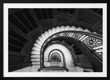 A mesmerizing black and white photograph shot from a high angle looking down into the center of a spiral staircase. The bright white marble steps contrast with the dark, ornate metal railing, creating a repeating arched pattern that draws the eye downward into a seemingly endless tunnel. The image is presented within a silver-colored frame. Artwork