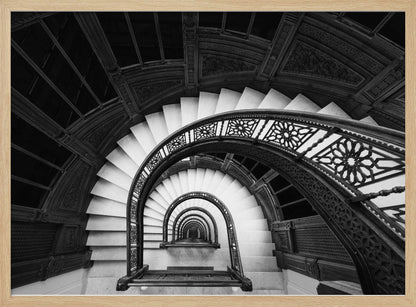 A mesmerizing black and white photograph shot from a high angle looking down into the center of a spiral staircase. The bright white marble steps contrast with the dark, ornate metal railing, creating a repeating arched pattern that draws the eye downward into a seemingly endless tunnel. The image is presented within a silver-colored frame. Artwork