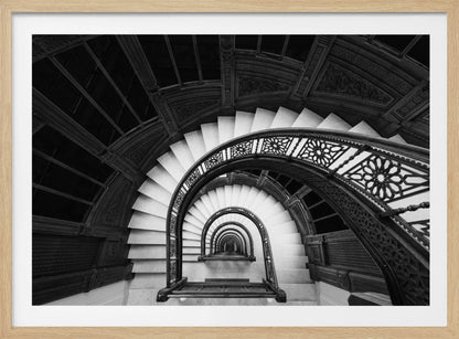 A mesmerizing black and white photograph shot from a high angle looking down into the center of a spiral staircase. The bright white marble steps contrast with the dark, ornate metal railing, creating a repeating arched pattern that draws the eye downward into a seemingly endless tunnel. The image is presented within a silver-colored frame. Artwork