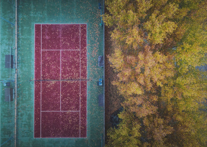 An aerial top-down photograph of a red and green tennis court covered in fallen leaves, adjacent to a dense grove of trees with vibrant yellow and gold autumn foliage, all enclosed in a silver frame. Wall Art