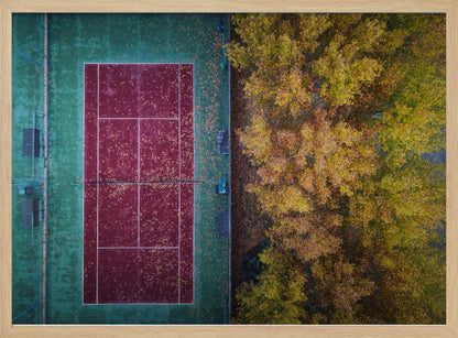 An aerial top-down photograph of a red and green tennis court covered in fallen leaves, adjacent to a dense grove of trees with vibrant yellow and gold autumn foliage, all enclosed in a silver frame. Wall Art