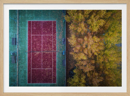 An aerial top-down photograph of a red and green tennis court covered in fallen leaves, adjacent to a dense grove of trees with vibrant yellow and gold autumn foliage, all enclosed in a silver frame. Wall Art