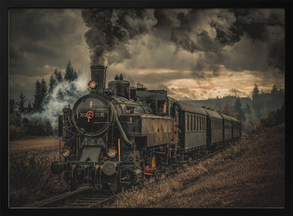 A powerful black steam locomotive on a railway track, billowing thick dark smoke into a dramatic, cloudy sky. The train travels through a rustic, grassy landscape, and the image has a warm, vintage color tone, all within a silver frame. Wall Art