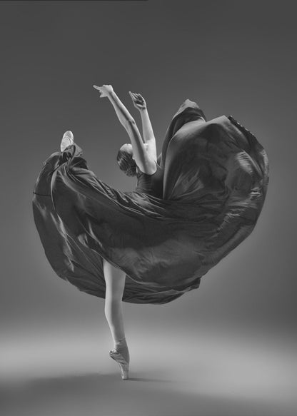 A dramatic black and white photograph of a ballerina captured mid-twirl. She is perfectly balanced on the tip of one pointe shoe, with her other leg kicked high. Her arms are raised elegantly above her head, and her long, dark dress billows out around her in a magnificent, fluid circle against a solid gray background. Wall Art