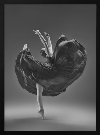 A dramatic black and white photograph of a ballerina captured mid-twirl. She is perfectly balanced on the tip of one pointe shoe, with her other leg kicked high. Her arms are raised elegantly above her head, and her long, dark dress billows out around her in a magnificent, fluid circle against a solid gray background. Wall Art