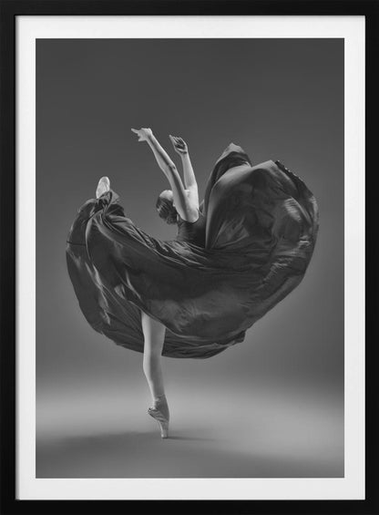 A dramatic black and white photograph of a ballerina captured mid-twirl. She is perfectly balanced on the tip of one pointe shoe, with her other leg kicked high. Her arms are raised elegantly above her head, and her long, dark dress billows out around her in a magnificent, fluid circle against a solid gray background. Wall Art