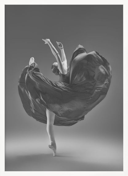 A dramatic black and white photograph of a ballerina captured mid-twirl. She is perfectly balanced on the tip of one pointe shoe, with her other leg kicked high. Her arms are raised elegantly above her head, and her long, dark dress billows out around her in a magnificent, fluid circle against a solid gray background. Wall Art