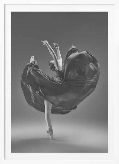 A dramatic black and white photograph of a ballerina captured mid-twirl. She is perfectly balanced on the tip of one pointe shoe, with her other leg kicked high. Her arms are raised elegantly above her head, and her long, dark dress billows out around her in a magnificent, fluid circle against a solid gray background. Wall Art