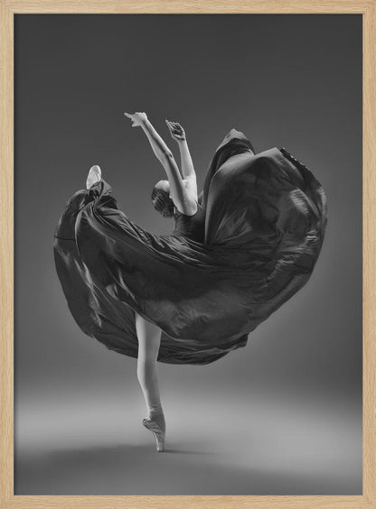 A dramatic black and white photograph of a ballerina captured mid-twirl. She is perfectly balanced on the tip of one pointe shoe, with her other leg kicked high. Her arms are raised elegantly above her head, and her long, dark dress billows out around her in a magnificent, fluid circle against a solid gray background. Wall Art