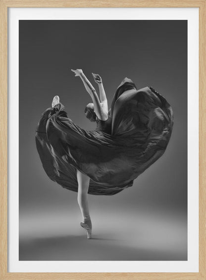 A dramatic black and white photograph of a ballerina captured mid-twirl. She is perfectly balanced on the tip of one pointe shoe, with her other leg kicked high. Her arms are raised elegantly above her head, and her long, dark dress billows out around her in a magnificent, fluid circle against a solid gray background. Wall Art