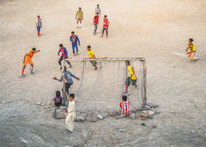An overhead view of a group of boys and young men playing a spirited game of soccer on a large, dusty dirt field. A player in a bright orange uniform prepares to kick the ball towards a makeshift goal, defended by a goalie in a dark blue jersey. Other players in various colored shirts are scattered across the field, while a few young spectators watch from the sidelines. The entire image is enclosed in a silver frame. Wall Art