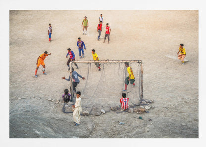 An overhead view of a group of boys and young men playing a spirited game of soccer on a large, dusty dirt field. A player in a bright orange uniform prepares to kick the ball towards a makeshift goal, defended by a goalie in a dark blue jersey. Other players in various colored shirts are scattered across the field, while a few young spectators watch from the sidelines. The entire image is enclosed in a silver frame. Wall Art