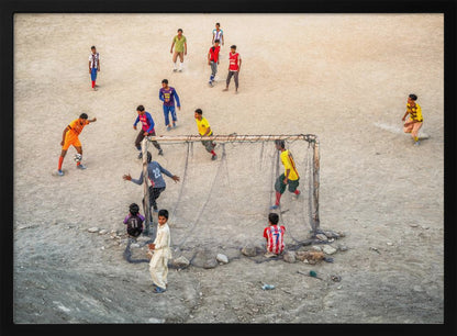An overhead view of a group of boys and young men playing a spirited game of soccer on a large, dusty dirt field. A player in a bright orange uniform prepares to kick the ball towards a makeshift goal, defended by a goalie in a dark blue jersey. Other players in various colored shirts are scattered across the field, while a few young spectators watch from the sidelines. The entire image is enclosed in a silver frame. Wall Art